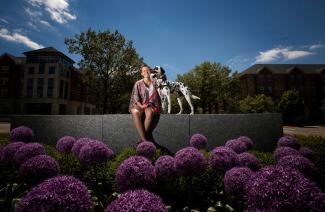 A student sits on a low wall behind a flower bed with a dog.